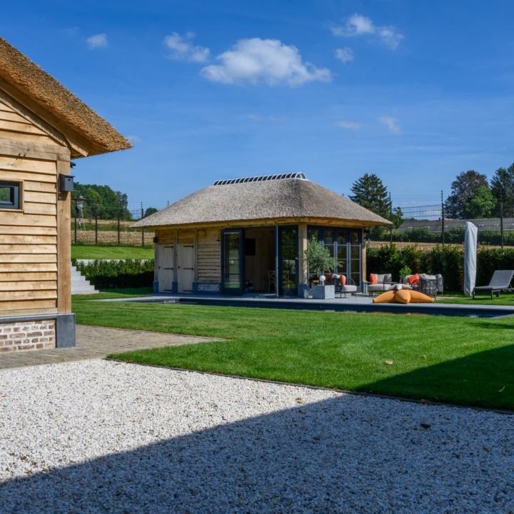 Uitzicht over tuin en zwembad richting vrijstaand houten poolhouse met rieten dak