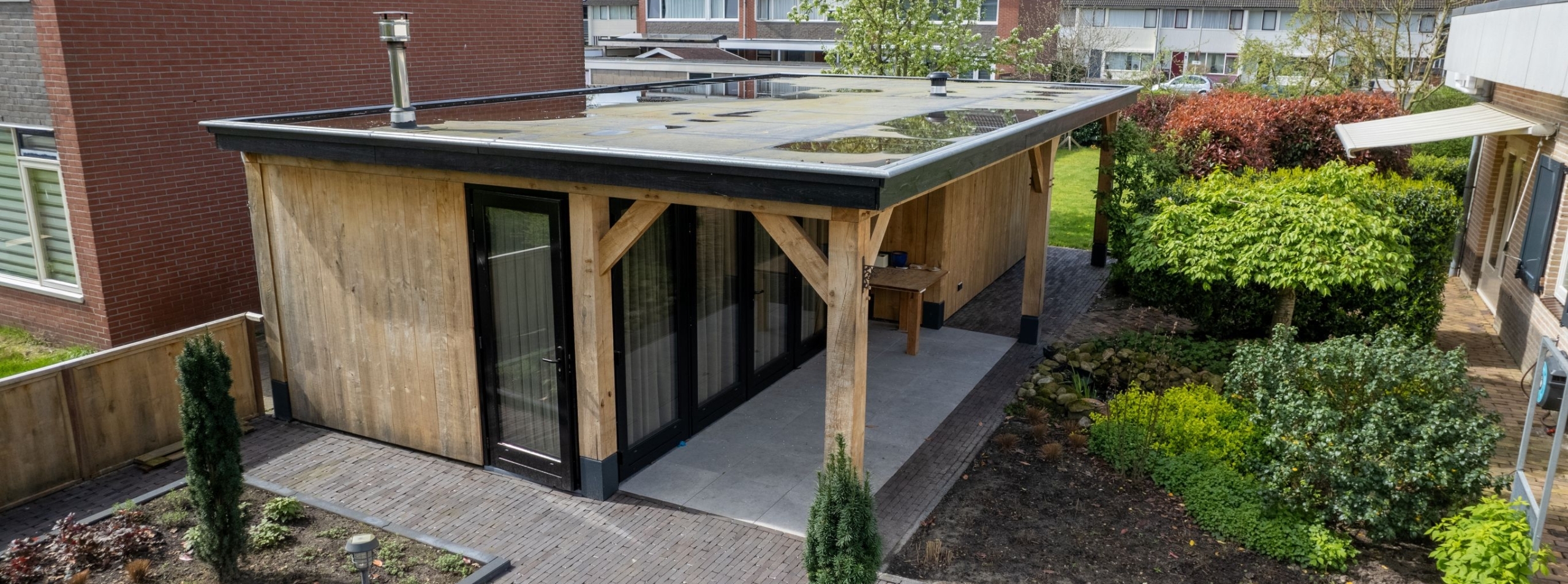 Eiken tuinhuis met plat dak, veranda en zwarte kozijnen in een aangelegde achtertuin